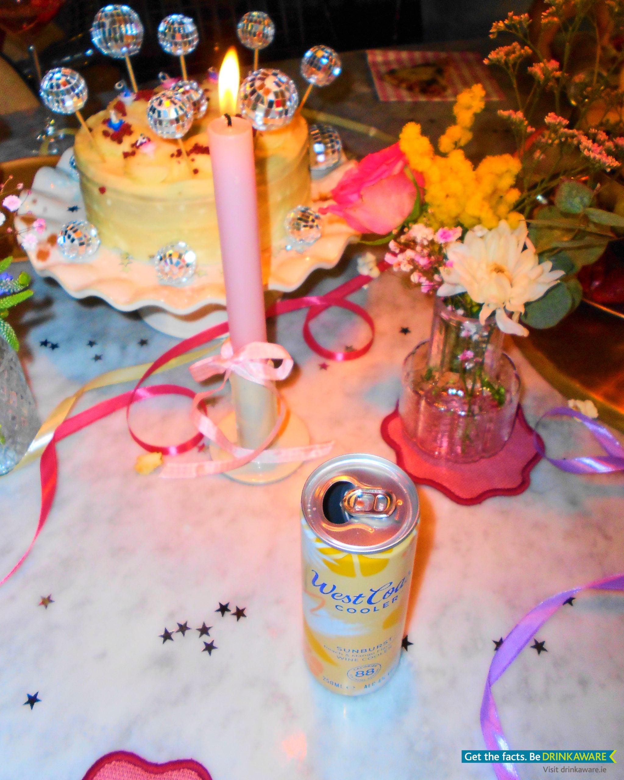 We see a can of West Coast Cooler Sunburst on a table decorated for a cute girly birthday party. There is cake in the background as well as candles and flowers.
