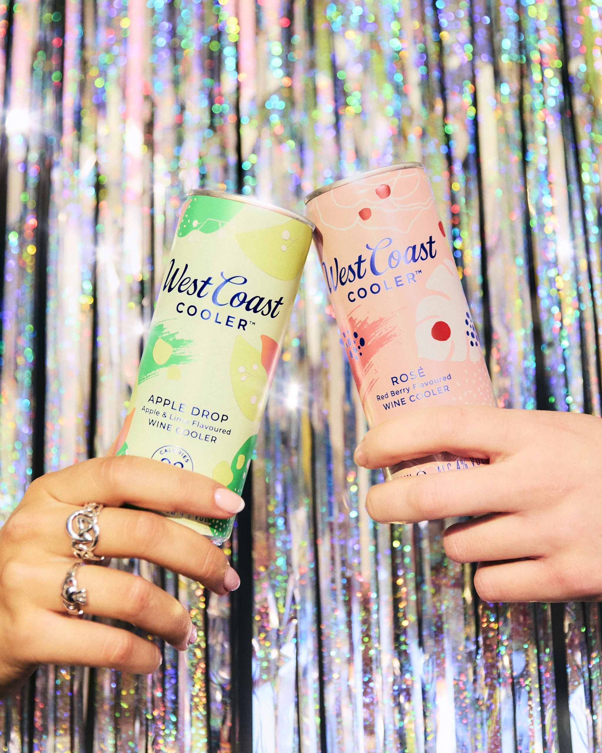 We see two hands, one holding a can of West Coast Cooler Apple Drop and the other a can of West Coast Cooler Rosé. They are cheersing in front of a silver glittery background